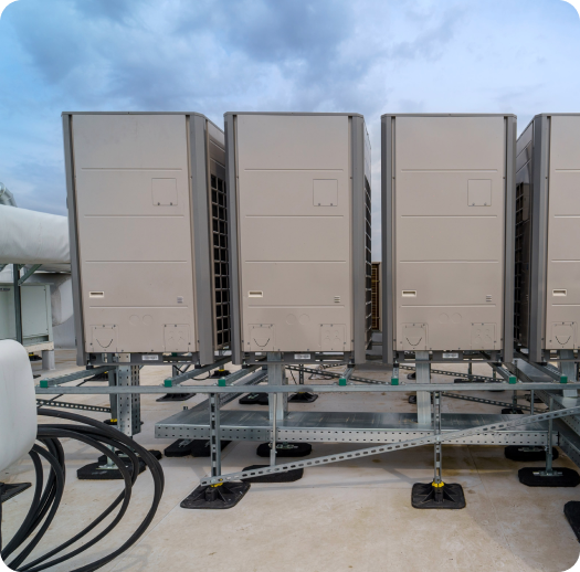 Four large industrial air conditioning units are installed on a rooftop under a cloudy sky, with connected pipes and cables visible around the equipment.