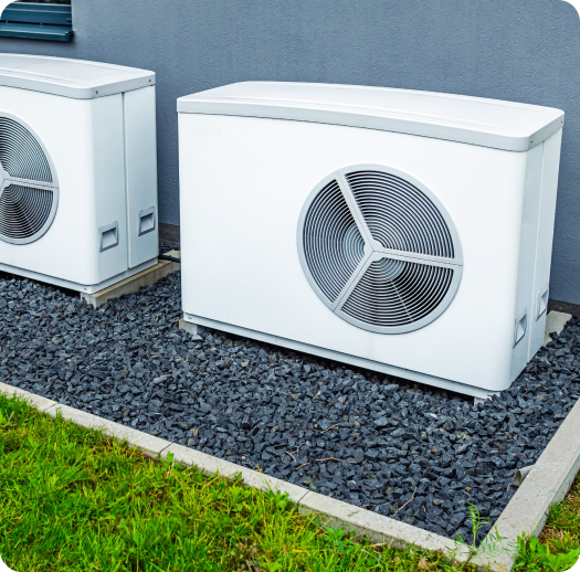 Two modern outdoor heat pump units with large circular fans are installed on a bed of black gravel next to a gray building wall. Grass is visible in the background.
