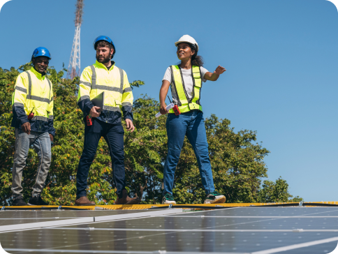 Three engineers in safety gear stand on a solar panel installation outdoors, with trees and a clear blue sky in the background. One person points ahead while the others look on.