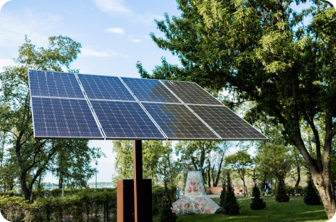 A large solar panel mounted on a metal stand is positioned outdoors on grass, surrounded by green trees and shrubs on a sunny day.
